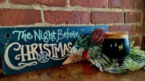 Image of a Christmas ale in a glass surrounded by holiday greenery next to a sign with the words The Night Before Christmas.