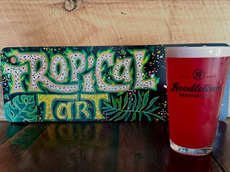 A pint glass of Hoodletown's tropical tart gose beer with a sign behind it featuring the beer name.